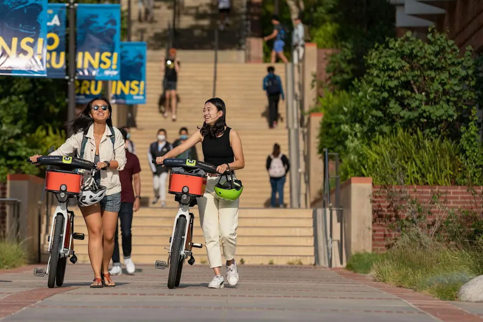 Two women walk with bikes on a sunny campus path.
