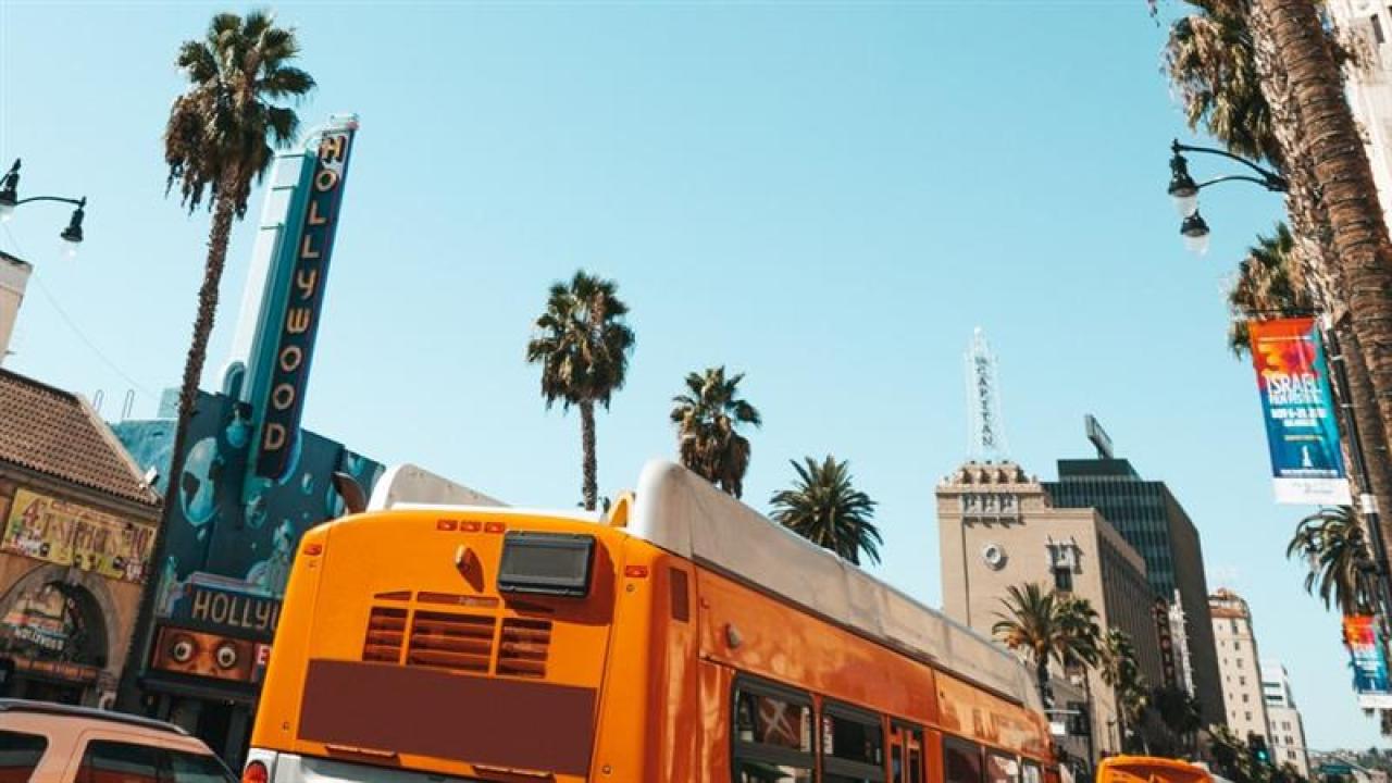 The image shows a vibrant street scene in Hollywood, California. Prominently featured is an orange public bus in the foreground, driving along a street lined with palm trees. In the background, there are iconic Hollywood landmarks, including a large vertical neon sign that reads "HOLLYWOOD." The sky is clear and blue, adding to the lively and sunny atmosphere typical of Los Angeles. Banners and other buildings also contribute to the bustling urban environment.
