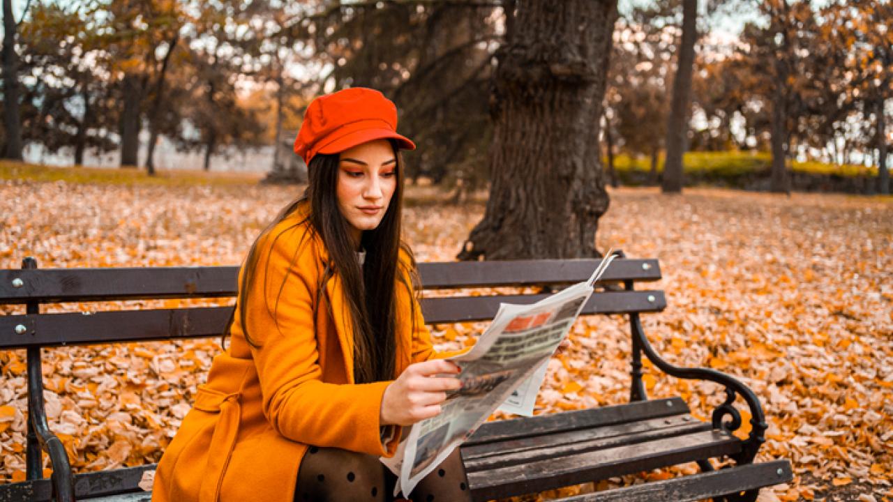 Woman dressed in orange reading a paper on a bench outside