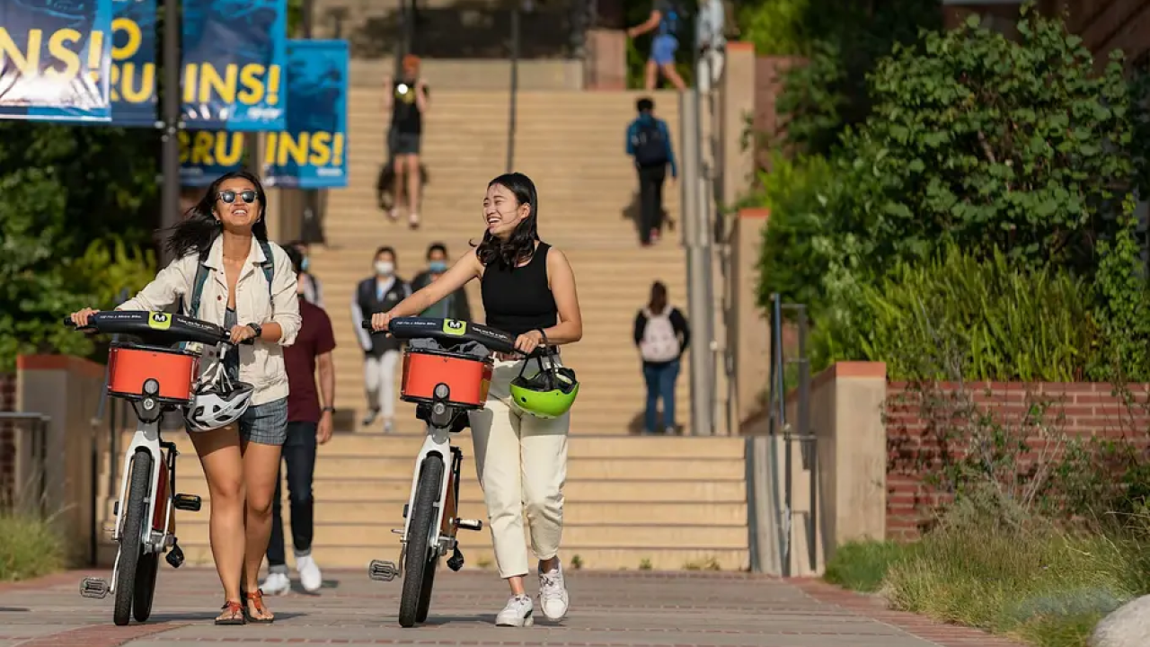 Two women walk with bikes on a sunny campus path.