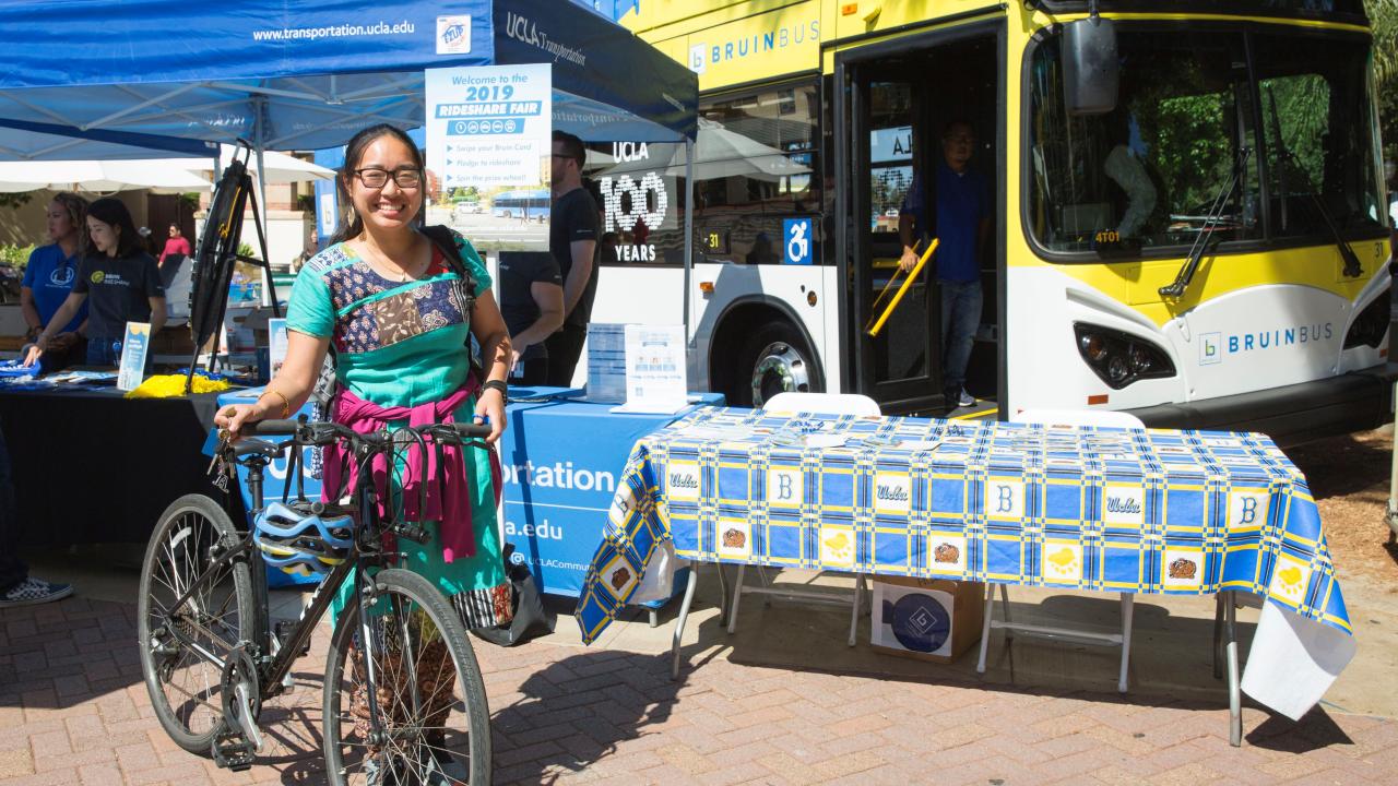 Female student with a bike at the Rideshare Fair with a BruinBus in the background