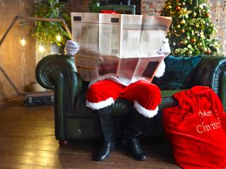 Santa reading the newspaper inside by the Christmas tree with his sack