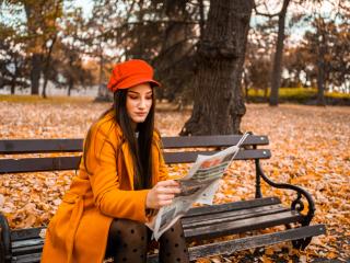 Woman dressed in orange reading a paper on a bench outside